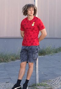 Rød grafisk T-shirt med korte ærmer og forskellige symboler, parret med mørke cargo-shorts og sorte sneakers, stående nær en grå pole.