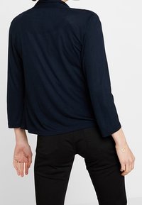 Marinblus, långärmad topp med avslappnad silhuett och rundade krage, med en mjuk textur och slät tyg. Svarta byxor synliga.