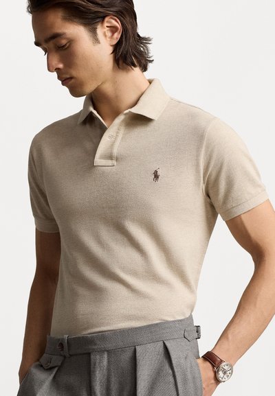 Beige polo skjorte i teksturert stoff med en tre-knapps placket og brodert logo; parret med grå pregede bukser og et brunt klokke.