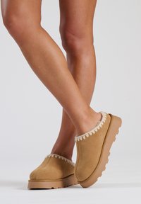Des chaussures slip-on couleur tan en suede doux, avec une bordure duveteuse et une semelle en caoutchouc texturée, dotées d'un design de plateforme basse.