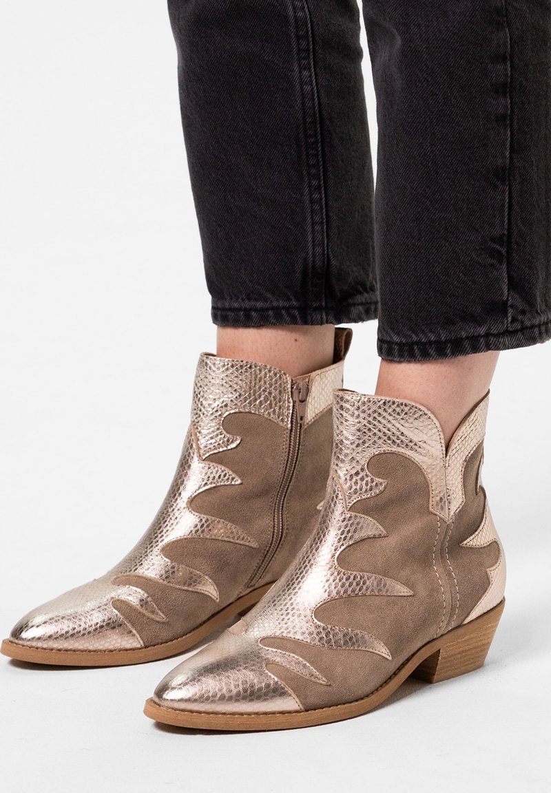 Stivali cowboy metallici oro e beige in pelle alti fino alla caviglia, con pannelli texture e lisci, indossati con jeans neri a vita bassa.
