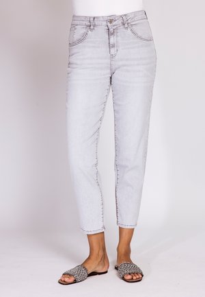 Hellblaue Jeans mit hoher Taille und knöchellanger Länge, getragen mit schwarz-weißen gewebten Slides bei einer Person vor einfarbigem Hintergrund.