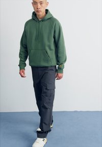 Haut à capuche en coton vert avec poche avant, associé à un pantalon cargo sombre avec poches latérales. Baskets blanches avec des accents jaunes.