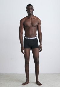 Svarte boxer-shortser med logo-midjebånd, laget av et glatt stoff. Modellen står mot en enkel bakgrunn og viser passformen.