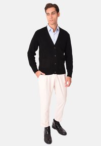 Cardigan nero a maglia con scollo a V, chiusura con bottoni e due tasche frontali, abbinato a pantaloni crema e stivali neri alla caviglia.