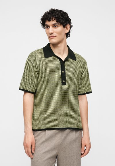 Νέος άνδρας με σγουρά μαλλιά που φοράει ένα πλεκτό πόλο με κοντά μανίκια σε κίτρινο και μαύρο με υφή και μπεζ παντελόνια, στέκεται μπροστά σε απλό φόντο.