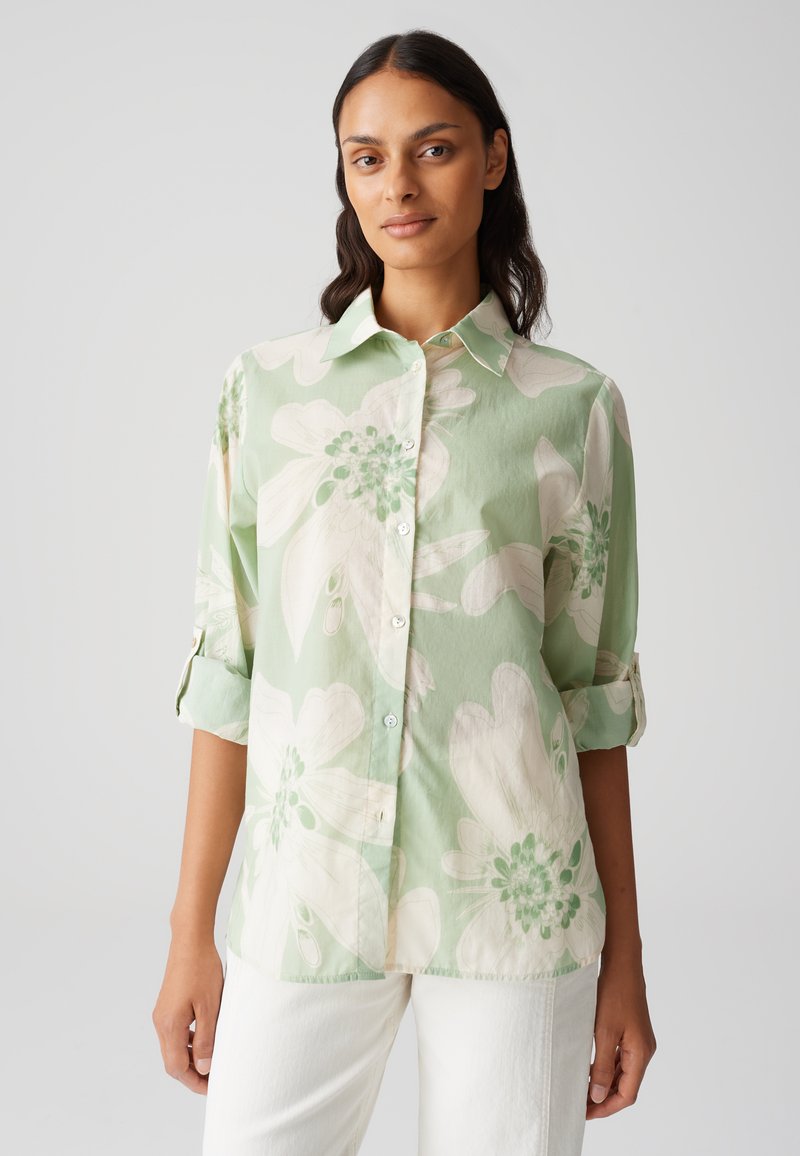 Camicia button-up verde chiaro con ampi motivi floreali bianchi, morbida al tatto e con maniche arrotolabili. Presenta colletto classico e chiusura anteriore con bottoni.