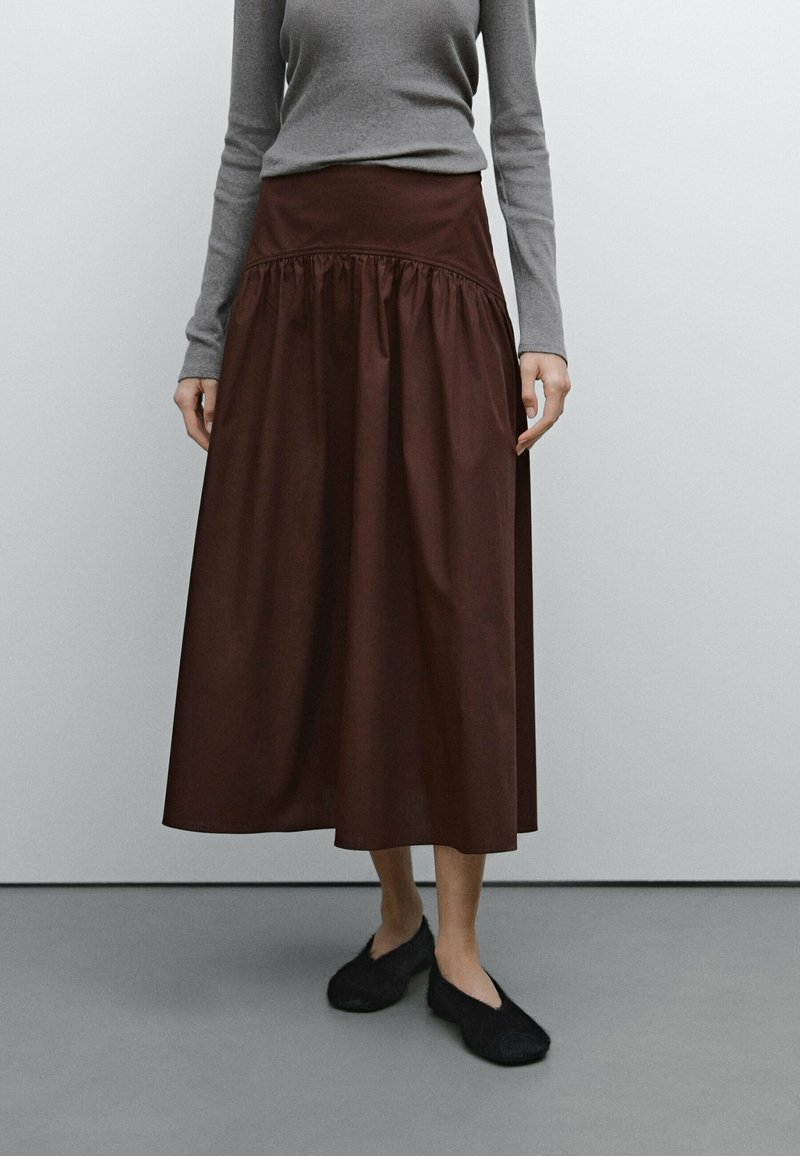 Longue jupe bordeaux à taille froncée, en tissu lisse. Associée à un haut gris à manches longues et des chaussures plates noires.