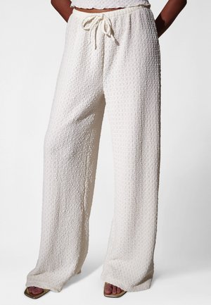 Pantalon blanc texturé à jambes larges avec taille à cordon, porté par une personne ayant les mains dans le dos, associé à des talons ouverts.