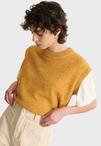Senfgelber gestrickter Pullover mit kurzen Ärmeln, ausgestattet mit einem gerippten Halsausschnitt. Über einem weißen Hemd getragen, kombiniert mit hellen Hosen.