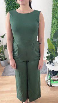 Nő áll bent egy ujjatlan sötétzöld overallban, fülbevalókkal és nyakláncokkal, növények és egy fényképezőgép és könyvek tálcájával körülvéve.