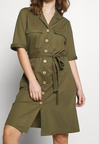 Robe chemise vert olive avec col, manches courtes, deux poches avant, ceinture à nouer à la taille et ourlet à hauteur du genou.