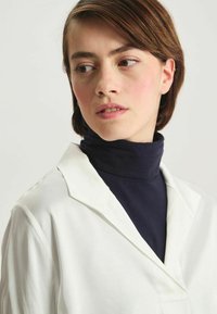 Blouse à col blanc portée sur un col roulé marine. Texture lisse, lignes épurées et design minimaliste axé sur la superposition.