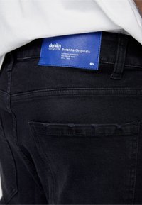 Svarta denimjeans med en blå etikett märkt "denim wear Bershka Originals," med minimalistisk sömnad och en slim, figurnära design.