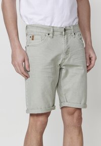 Moški v svetlo sivih zavihanih jeans kratkih hlačah in belem majici, stoji z rokami sproščeno ob straneh.