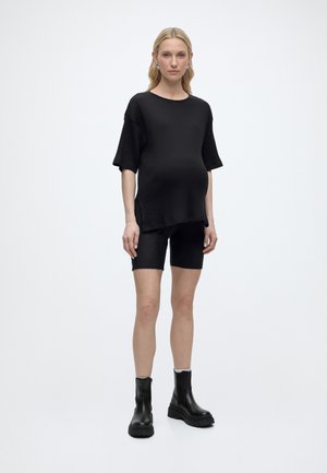 Femme portant un t-shirt noir côtelé ample, un short assorti longueur genou, des bottines noires, debout sur un fond blanc uni.