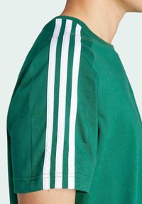 Zaļš kokvilnas t-krekls ar baltām trīs joslu detaļām uz pleca, īsām piedurknēm un apaļo kakla izgriezumu.