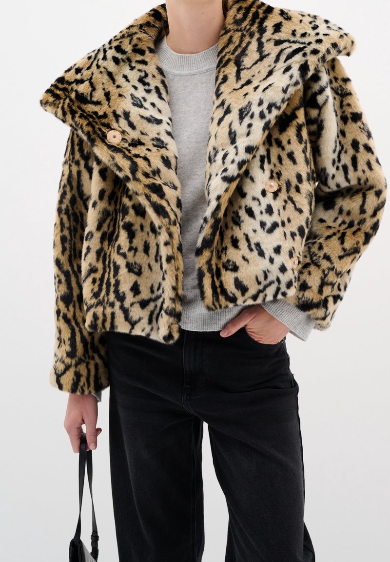 Veste en fausse fourrure imprimé léopard avec large col, motifs beige et noir, deux boutons, portée sur un pull gris et un jean noir.