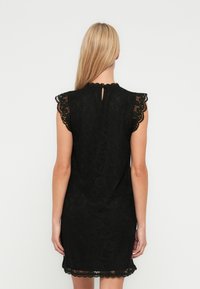 Vestido de renda preta com mangas curtas com acabamento em ondas, detalhe nas costas em forma de fechadura e silhueta reta com barra também acabada em ondas.