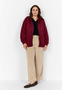 Sudadera burdeos con cremallera y capucha, combinada con pantalones de pana beige anchos. Los pantalones tienen rayas verticales y el calzado cuenta con tachuelas doradas.