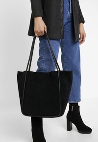 Svart mocka toteväska med läderdetaljer, vid öppen form, bärs av en person i en grå kappa och blå jeans, med svarta klackade ankelstövlar.