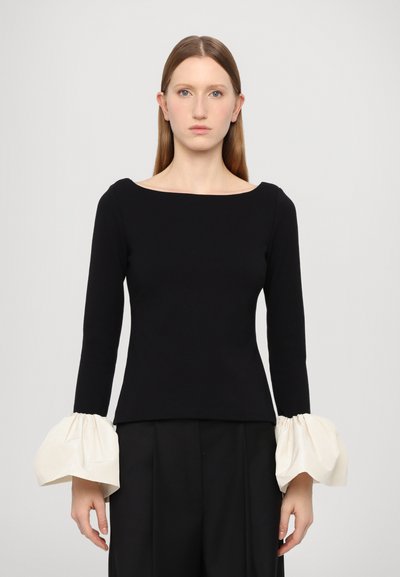 Haut noir à manches longues avec un large col et un tissu crème volumineux aux poignets. Texture lisse, coupe ajustée et design minimaliste.
