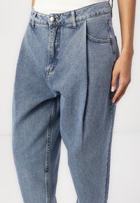 Højhængte blå denim jeans med plisseret front, udstyret med en sølvknap og to sidelommer, lavet af blødt, struktureret bomuldsstof.