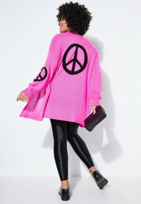 Jasně růžový oversize svetr s černými symboly míru na rukávech a zádech, doplněný černými lesklými legínami a botami.