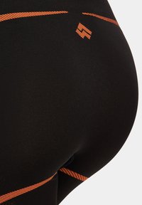 Czarne legginsy sportowe z pomarańczowymi paskami i logo. Materiał wydaje się gładki i elastyczny, zaprojektowany z myślą o komforcie i swobodzie ruchów.