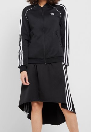 Person trägt eine schwarze Adidas Trainingsjacke und einen passenden Rock mit weißen Streifen an Ärmeln und Seiten, steht vor einem schlichten Hintergrund.
