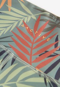 Abbigliamento sportivo stampato con sfondo verde e motivi di foglie rosse e blu scuro. Il nome del marchio "ACTIVE" è impresso in oro.