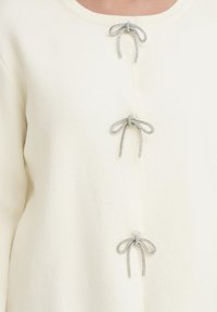 Pulover v kremni barvi z tremi okrasnimi srebrnimi dugimi zapiranji v obliki mašne, z gladko, mehko teksturo in preprostim okroglim izrezom.