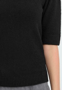 Sort strikket sweater med korte ærmer, der udviser en blød tekstur og ribbet kant. Materialet virker let og hyggeligt.