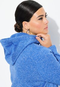 Blå huvtröja med en texturerad, melerad mönster, med huva och ribbade mudd. Modellen rör vid kragen och har på sig silverfärgade hoopörhängen.