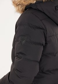 Sort puffjakke med hætte, der har en betræk af faux pels, quiltet struktur og et mærke på overarmen.