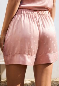 Pantaloni scurți roz din satin cu talie elastică, cu un model cu inimioare și o margine la tiv. Textură netedă, croială lejeră.
