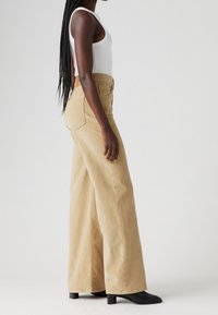 Jeans de pierna ancha beige de denim, de cintura alta, con bolsillos frontales y un bolsillo trasero cosido. Botas negras de tacón completan el look.