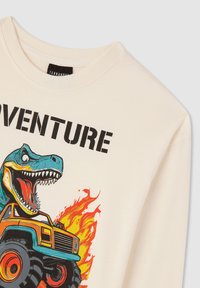 Kremowa koszulka z długim rękawem z grafiką niebieskiego dinozaura i pomarańczowego monster trucka z płomieniami. Gruby czarny napis głosi "PRZYGODA."