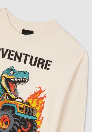 T-shirt a maniche lunghe color crema con una grafica di un dinosauro blu e un camion mostruoso arancione con fiamme. Testo in grassetto nero che recita "AVVENTURA."
