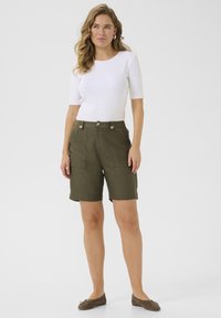 Haut à manches courtes blanc à côtes et short taille haute vert olive avec deux poches avant et boutons dorés. Des ballerines marron complètent la tenue.