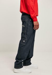 Dark denim cargo pants with multiple pockets, contrasting yellow stitching, and a relaxed fit. Paired with white sneakers and a red long-sleeve shirt.