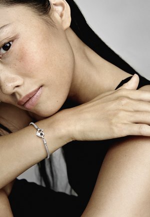 Bracciale d'argento con una chiusura a forma di cuore, caratterizzato da un design a catena intrecciata. La modella appoggia delicatamente la mano sul braccio, mettendo in mostra il gioiello.