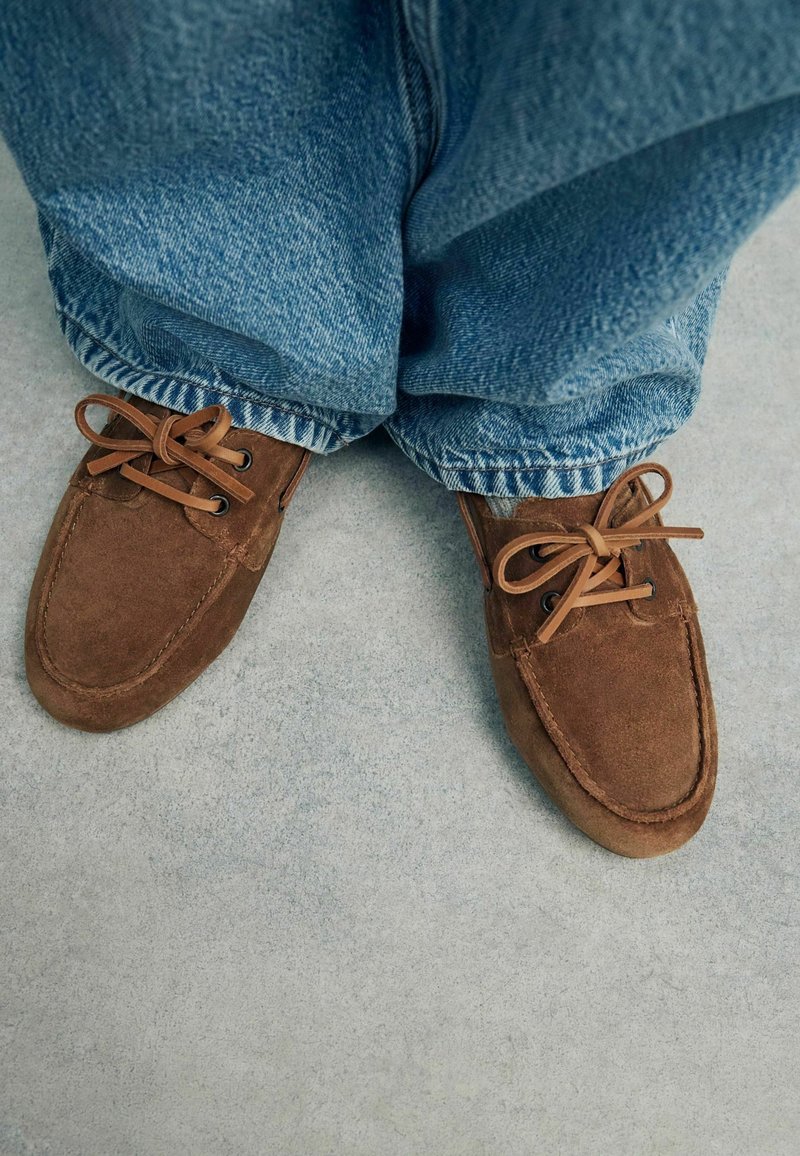 Chaussures marron en daim à lacets portées avec un jean en denim bleu, debout sur un sol en béton gris.