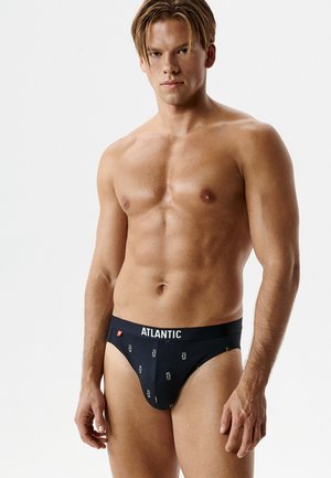 Muskularny mężczyzna z krótkimi brązowymi włosami w czarnych slipach marki Atlantic, na jasnym jednolitym tle, zwrócony przodem, z neutralnym wyrazem twarzy.