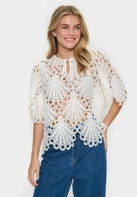Top bianco in pizzo con elaborate aperture decorative, scollatura a chiave e maniche corte svasate, abbinato a jeans blu a vita alta.