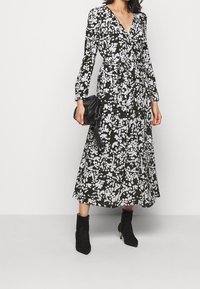 Musta kietaisumekko, jossa valkoinen kukkakuvio, pitkät hihat, solmittava vyötärö ja keskipitkä helma, yhdistetty mustiin nilkkureihin ja pikkulaukkuun.