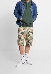 Jeanjacka över en mörkgrön hoodie, kombinerad med kamouflagemönstrade cargoshorts. Vita sneakers med svarta detaljer och vita strumpor fulländar stilen.