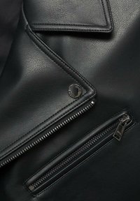 Veste en cuir noir avec finition texturée, présentant une poche à rabat angulaire, des fermetures éclair en argent et un détail de bouton-pression rond avec logo.