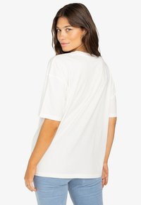 Camisa blanca de gran tamaño con mangas cortas, cuello redondo y tela suave, combinada con pantalones acanalados azul claro, vista desde atrás.