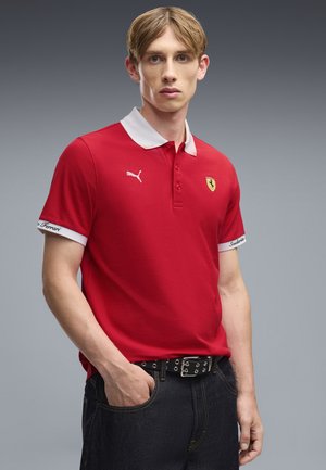 Młody mężczyzna o blond włosach, ubrany w czerwoną koszulkę polo Puma Ferrari i ciemne jeansy, stojący z jedną ręką w kieszeni.